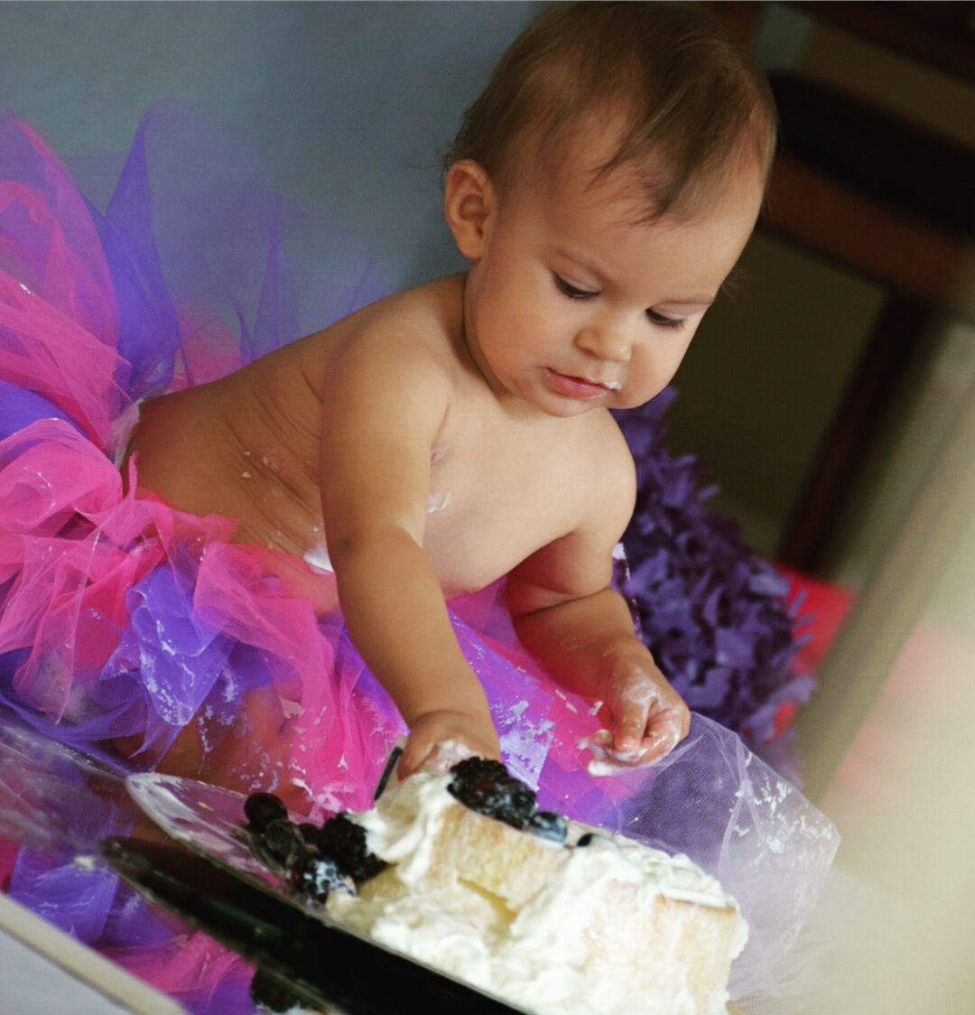 A baby in a pink and purple tutu reaching into a smash cake