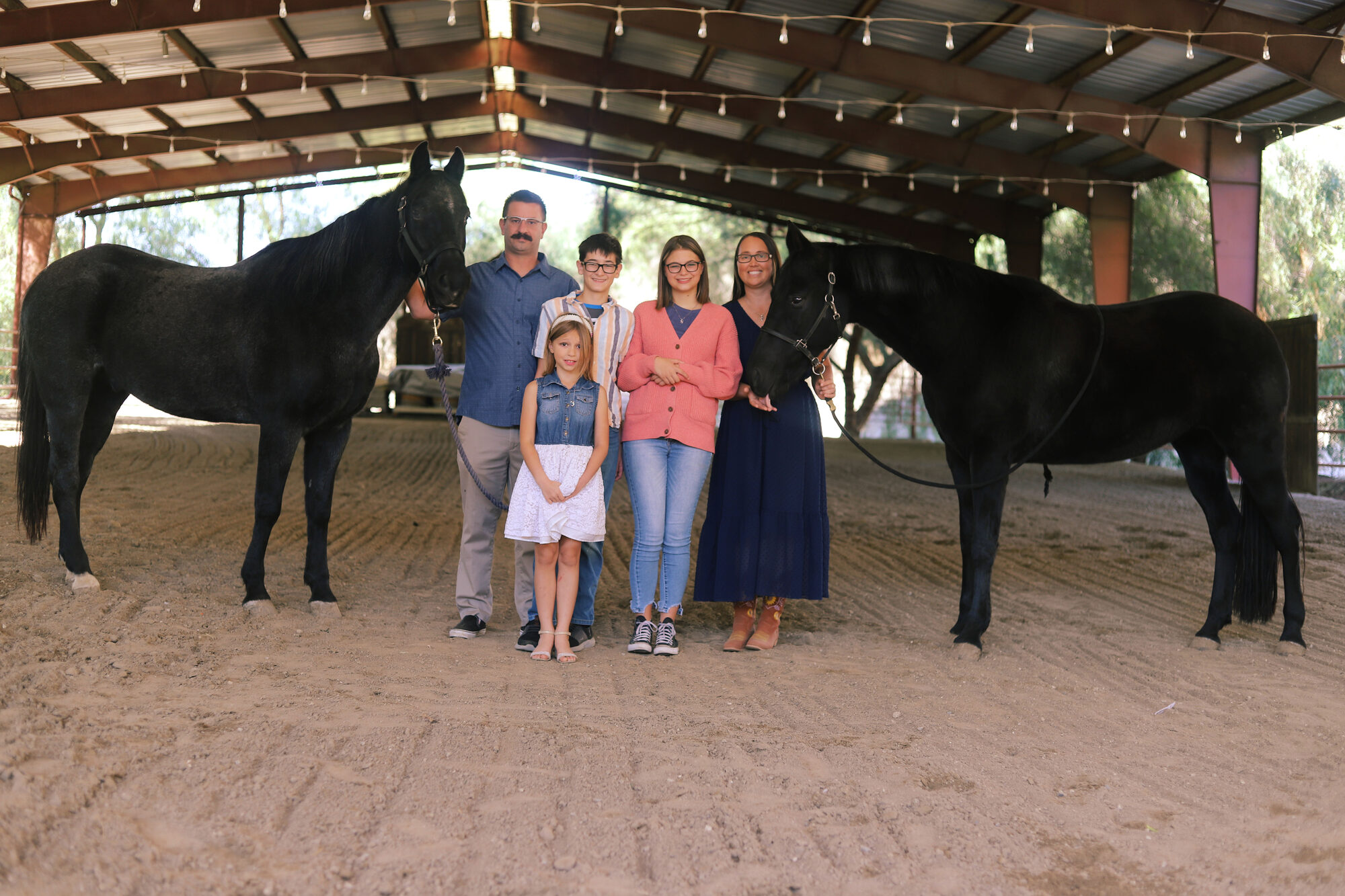 A family of five standing between two black horses in a covered arena