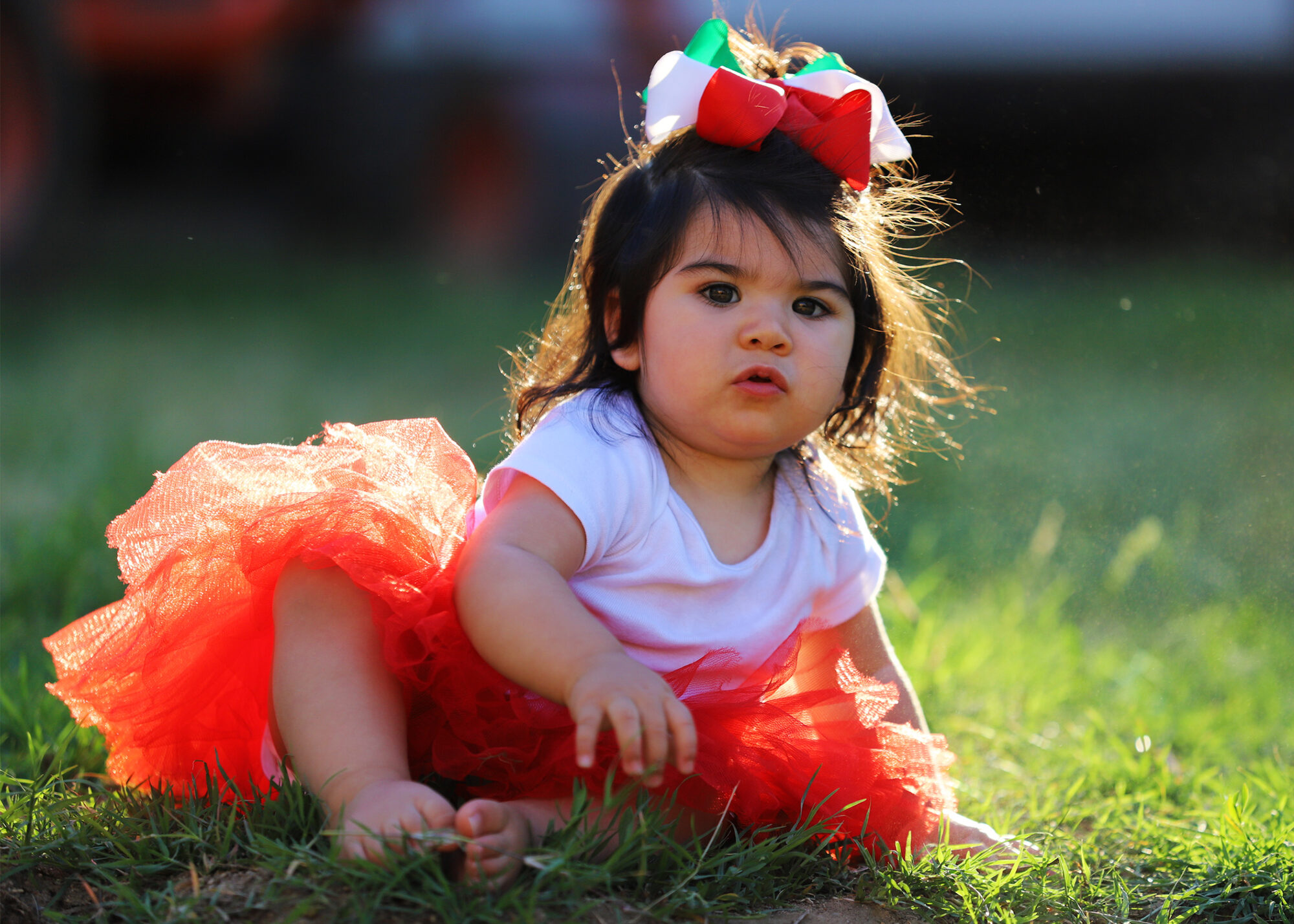 A toddler in a red tutu and hair bow sitting on the grass